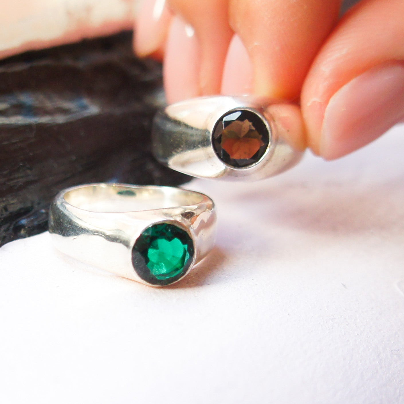 Silver rings with green and black gemstones held by a hand on a white surface.