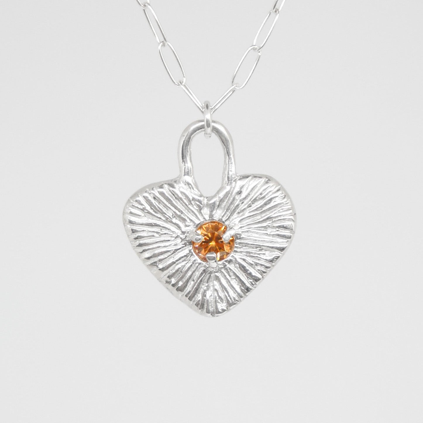 Silver heart-shaped pendant with a small orange gemstone on a white background