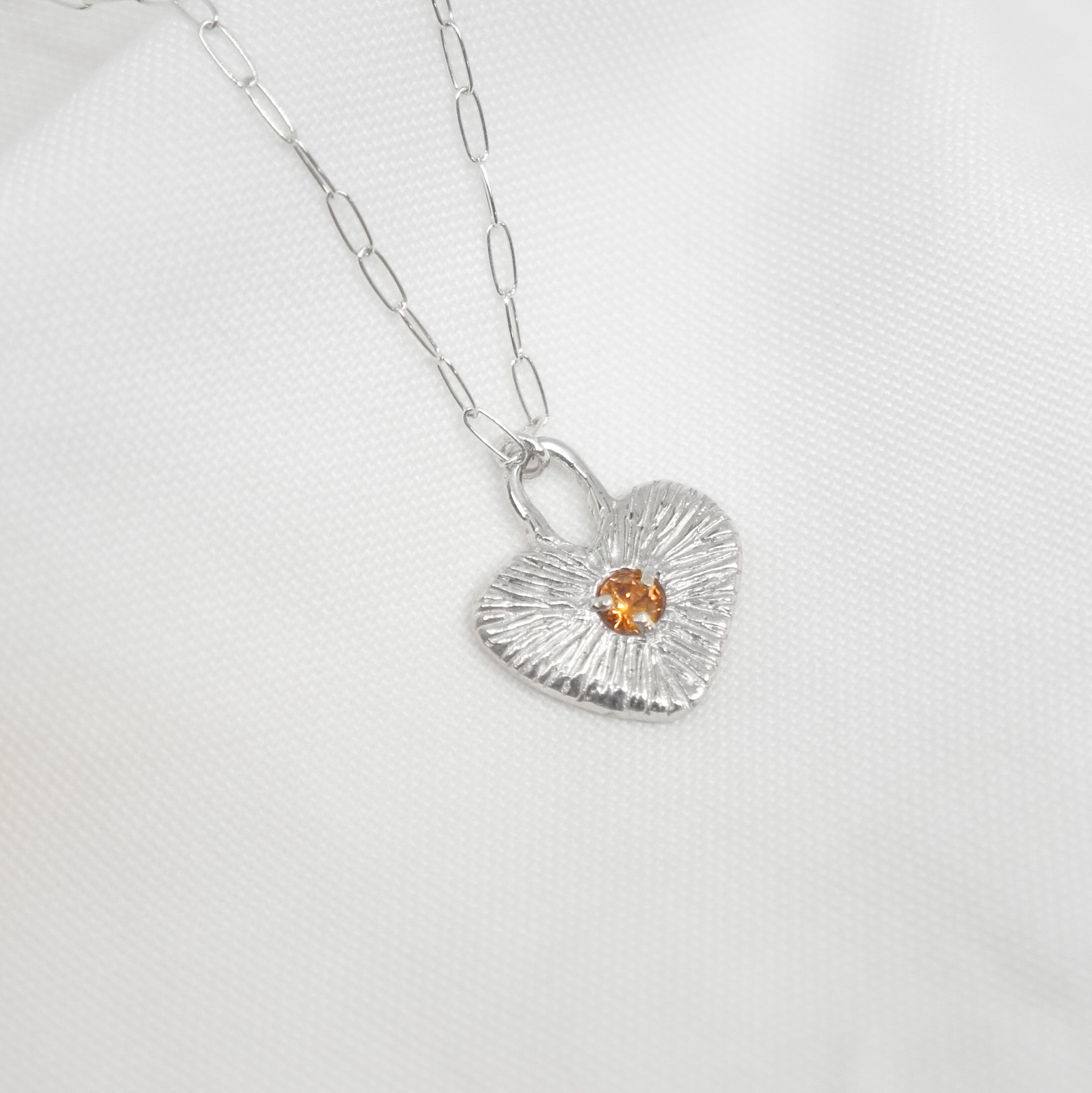 Silver necklace with a heart-shaped pendant featuring a small orange gemstone on a white background