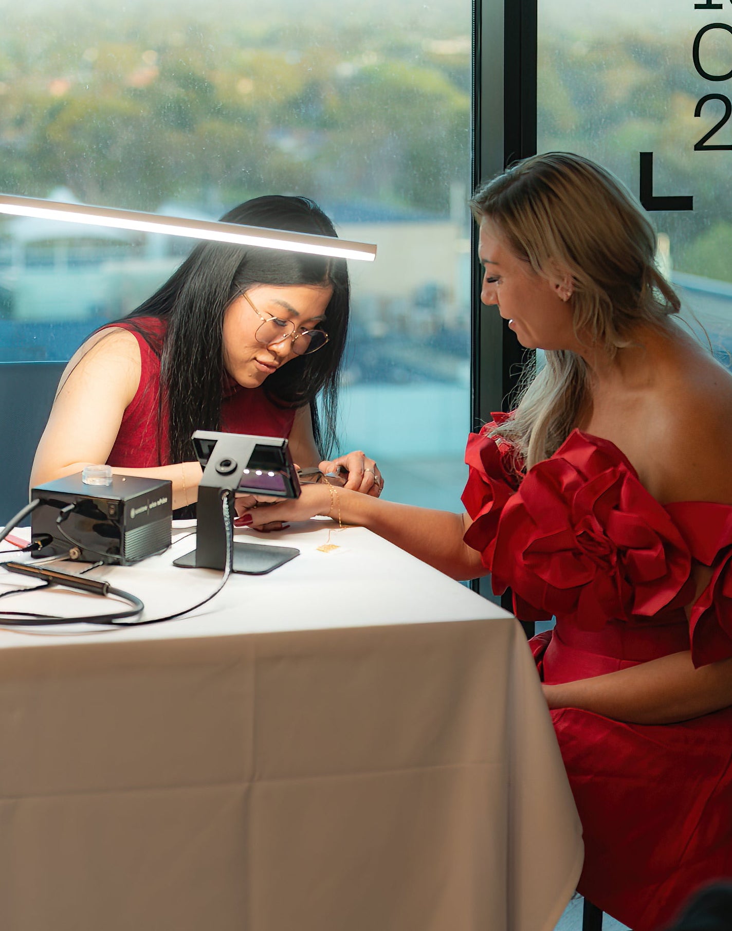 Two women in red dresses sitting at a table with welding Permanent Jewellery