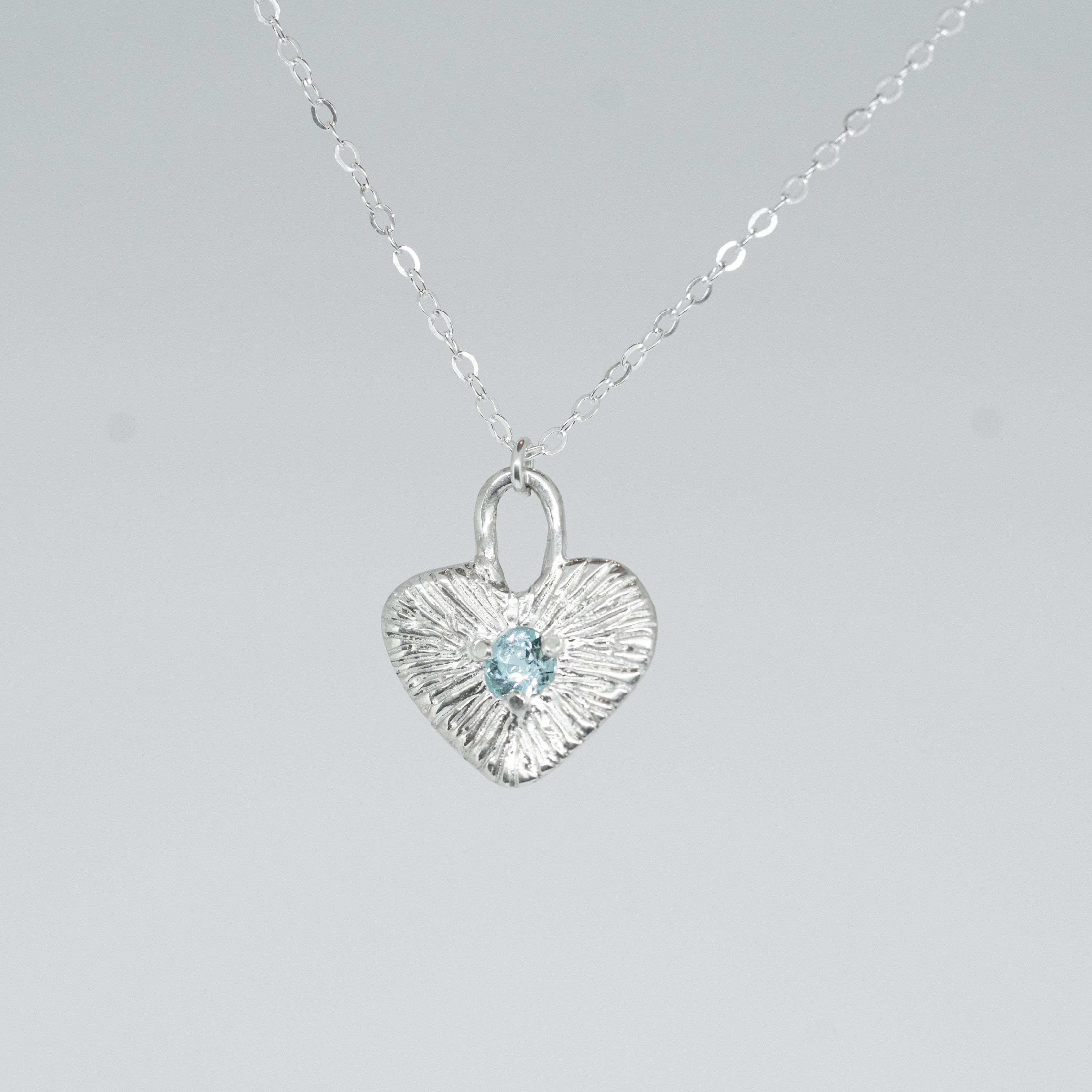 Heart-shaped silver pendant with a blue gemstone on a gray background